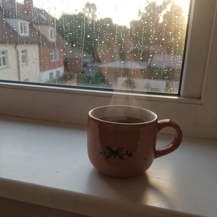 A ceramic coffee cup steaming on a windowsill with rain on the glass behind it.